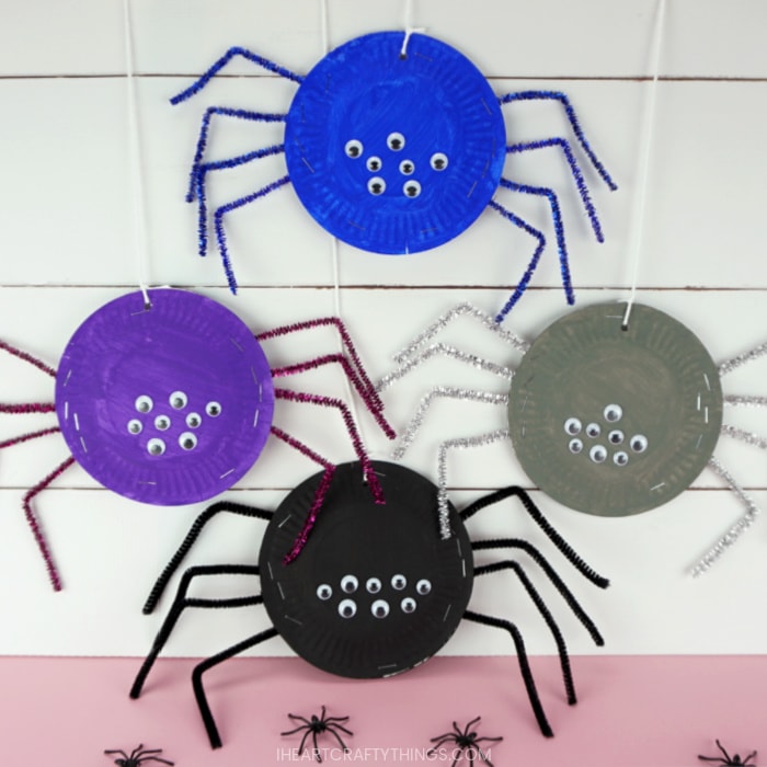 A blue, purple, gray and black paper plates spider hanging from a string on a white shiplap background.