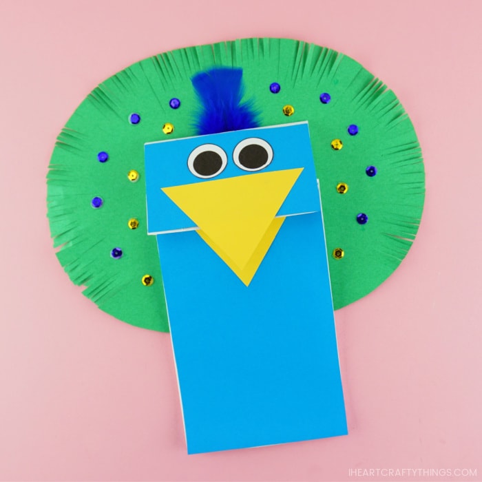 Paper bag peacock puppet craft laying slightly at an angle on a pink background.