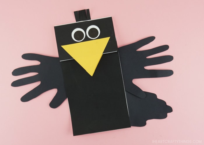 Paper bag crow puppet finished and laying flat on a pink background.