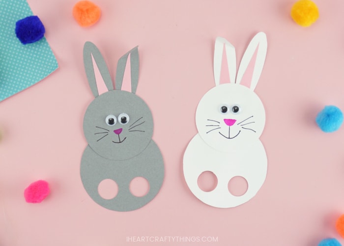 A gray bunny puppet and a white bunny puppet laying next to each other on a pink background with colored poms scattered around.
