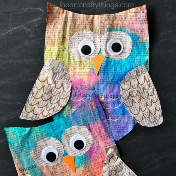 Two painted newspaper owl crafts, one laying on top of the other on a black chalkboard background.