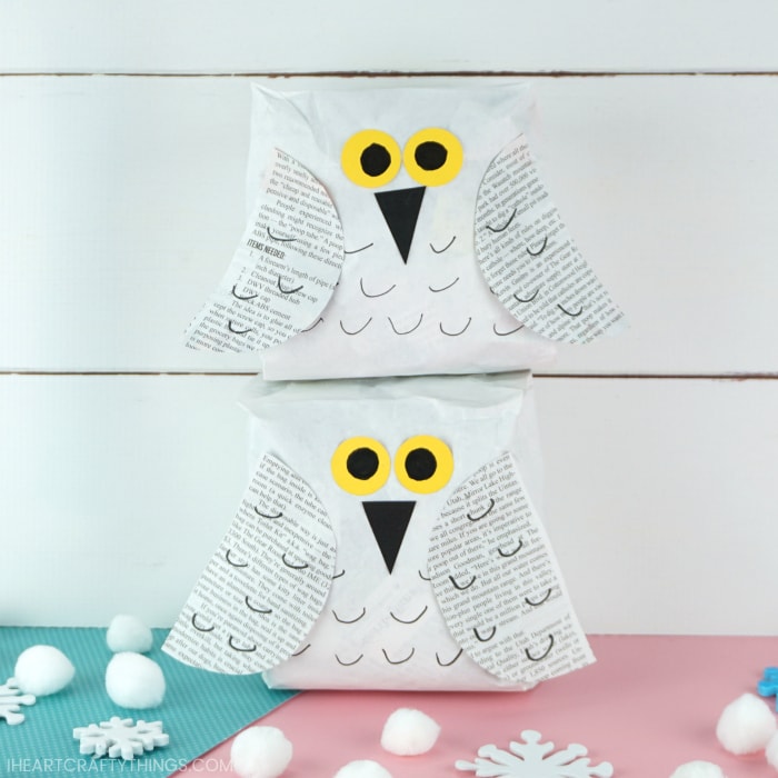 Two snowy owl crafts, one laying on top of the other in front of a white shiplap background with white pom poms scattered around the bottom.