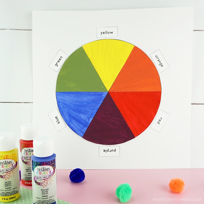 Finished color wheel craft propped against a white shiplap background with Testors red, yellow and blue acrylic paint bottles and craft poms sitting in front of the artwork.