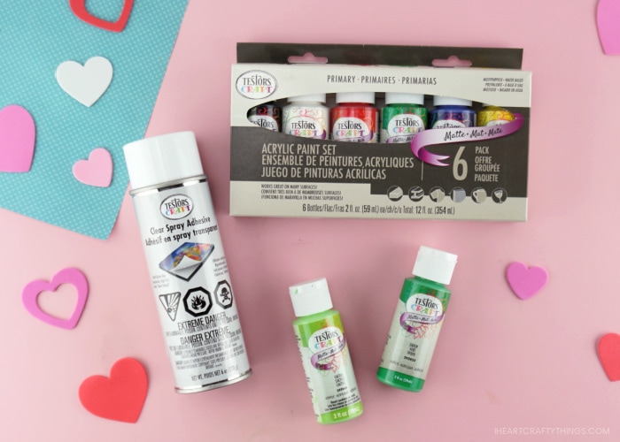 Assortment of Testors Craft acrylic paints and Testors spray adhesive laying down on a pink background with heart stickers scattered around.