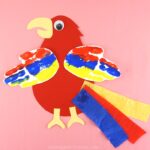 red, blue and yellow handprint parrot craft laying flat on a solid pink background.