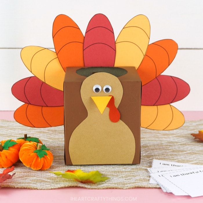 Close up square image of a tissue box made into a thankful turkey box sitting on a brown table runner with mini felt pumpkins, some fake leaves and the thankful writing note cards scattered around.