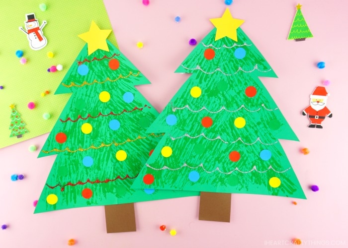 Two fork painted Christmas tree crafts laying side by side on a pink background with colorful pom poms and stickers scattered around.