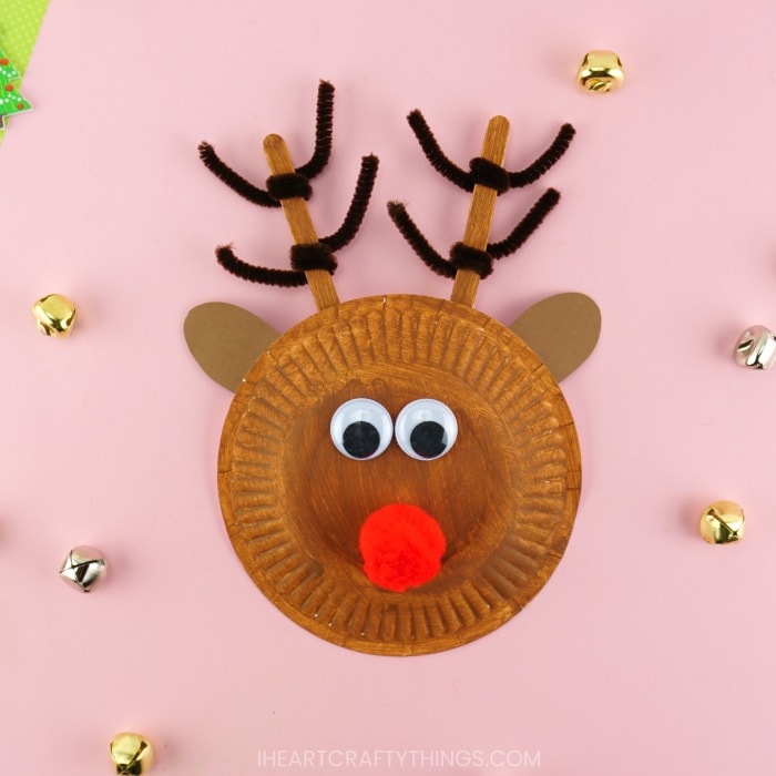 Square image of completed paper plate reindeer craft laying on a pink background with bells scattered around.