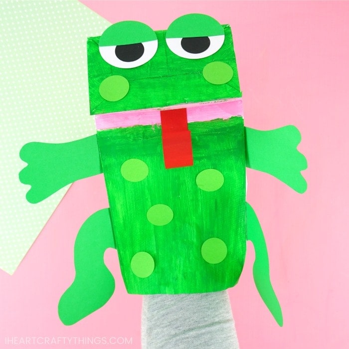 Someone with their hand inside the paper bag frog puppet on a pink background