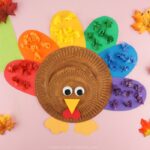 Square close up image of rainbow turkey craft laying on a pink background with fall colored leaves scattered around the turkey.