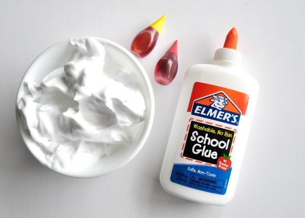 Bowl of shaving cream laying next to red and yellow food coloring and bottle of Elmer's school glue.