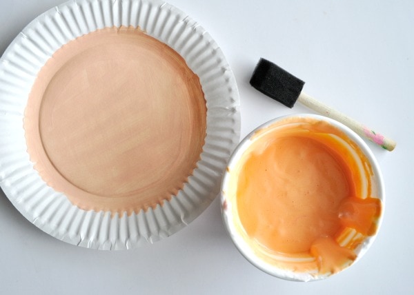 White paper plate with the center circle painted with peach paint sitting next to a bowl of mixed orange puffy paint and a paintbrush ready to paint the paper plate with.