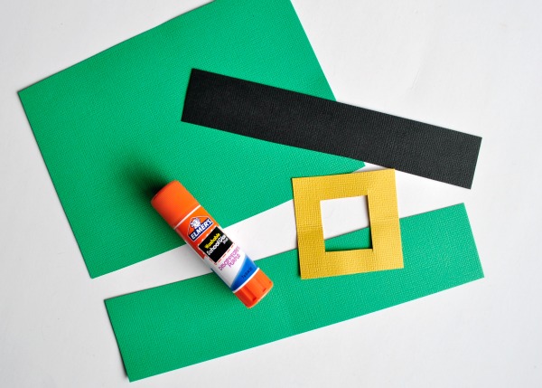 Leprechaun hat pieces cut out of green black and yellow cardstock paper with a Elmer's glue stick sitting on top of them.