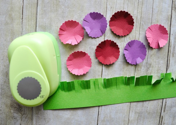 pink, purple and red scallop circles cup out laying next to a scallop circle punch and a rectangle of green cardstock paper that has slits cut along one end to look like blades of grass.