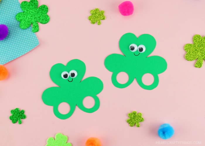 Two shamrock finger puppets laying side by side on a pink background with glittery shamrock stickers and colored poms scattered around them.