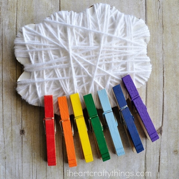 Square image of yarn wrapped cloud and rainbow painted clothespins attached to it sitting on a faux wood background.