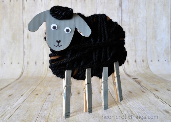 Vertical image of yarn wrapped sheep craft with clothespin legs standing on faux wood background.