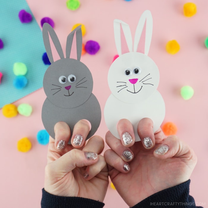 Adults with their fingers in two bunny finger puppets, one white bunny and one gray bunny, with a pink out of focus background with colored poms scattered around.