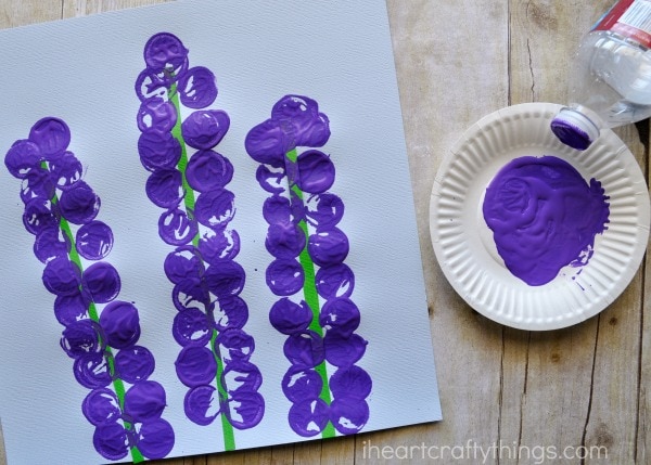 Small paper plate with purple paint on it and a plastic water bottle laying next to it, showing how to the dip the cap into the purple paint and onto the paper to make the hyacinth flower petals.