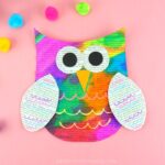 Painted newspaper owl craft laying on a pink background with craft pom poms scattered around.