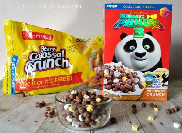 Box of Kung Fu Panda cereal and a bag of Berry Colossal Crunch cereal with a bowl full of cereal and some scattered around a faux wood background.