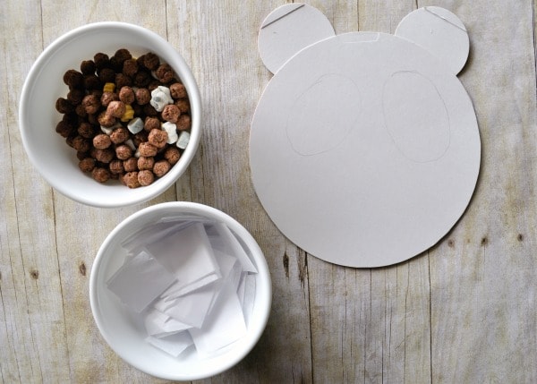 Cereal box panda shape laying on a faux wood background with a bowl of white tissue paper and cereal ready to make the craft.