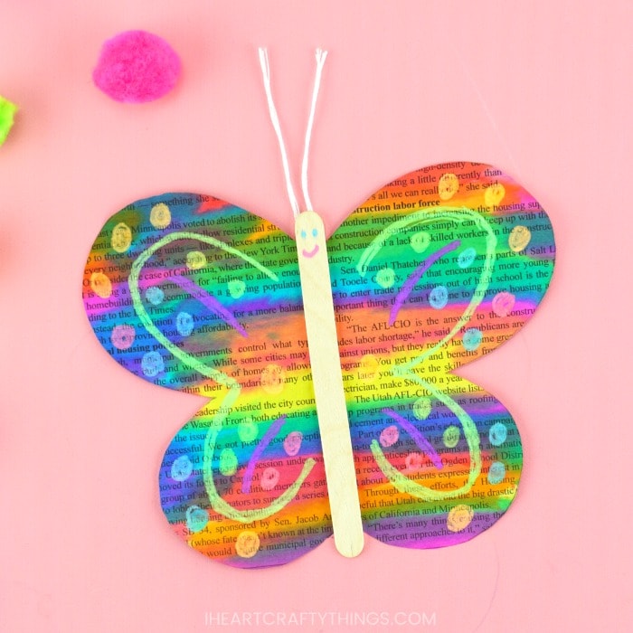 Close up square image of painted newspaper butterfly craft laying on a pink background with a pink pom pom scattered in the background.