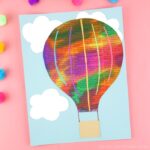 Painted newspaper hot air balloon craft laying on a pink background with craft poms scattered around.