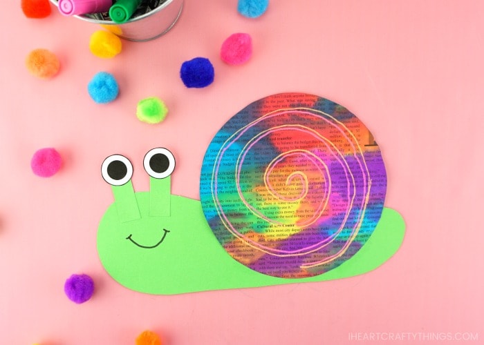 Horizontal image of finished newspaper snail craft laying on a pink background with colored craft pom poms scattered around in the background.