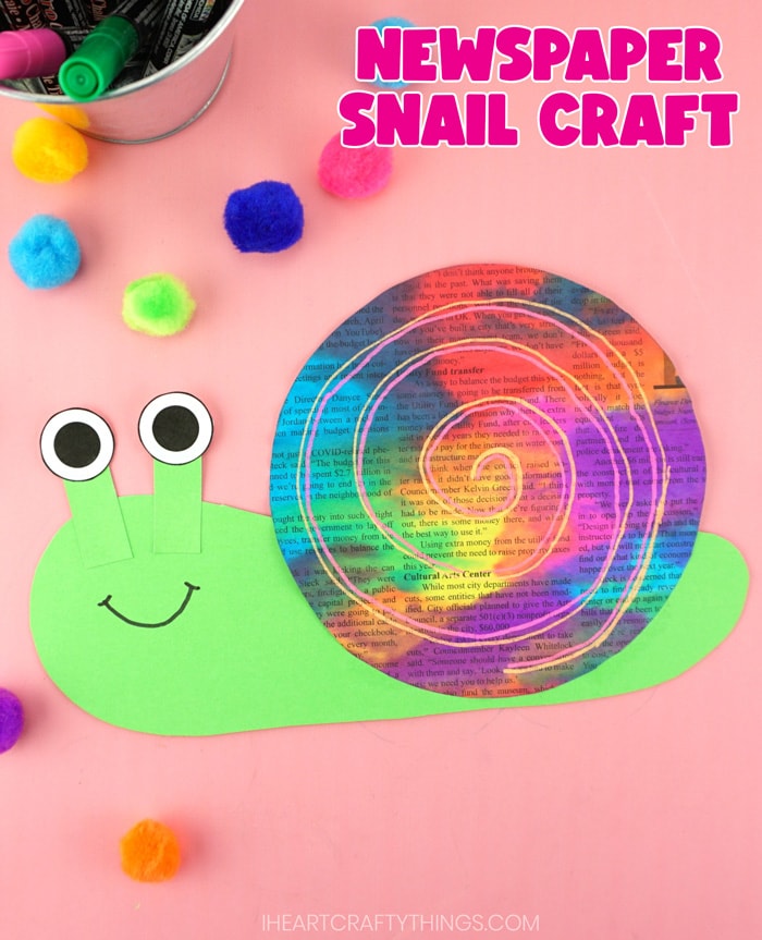 Vertical close up image of finished snail craft laying on a pink background with colored craft poms scattered around and metal tin of chalk markers in the top left corner. Words "Newspaper snail craft" are in the top left corner.