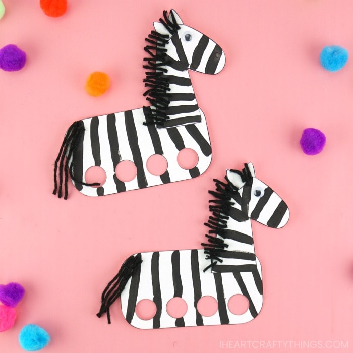 Two zebra finger puppet crafts laying next to each other on a pink background with colored craft pom poms laying around them.