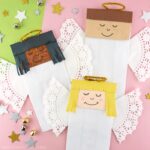 Three paper bag angel crafts laying at different angles on a pink background with star stickers and bells scattered around.