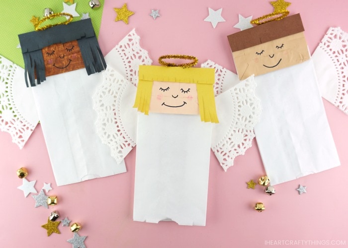 Horizontal image of three paper bag angel crafts laying next to each other on a pink background with stars and bells scattered around.