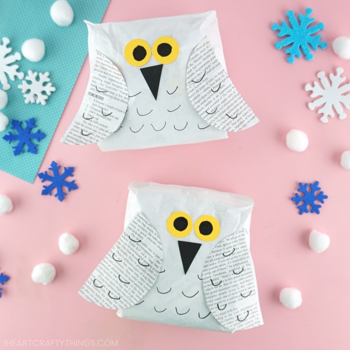 Two snowy owl crafts made from paper bags laying face up on a pink background with snowflake stickers and white pom poms scattered around.