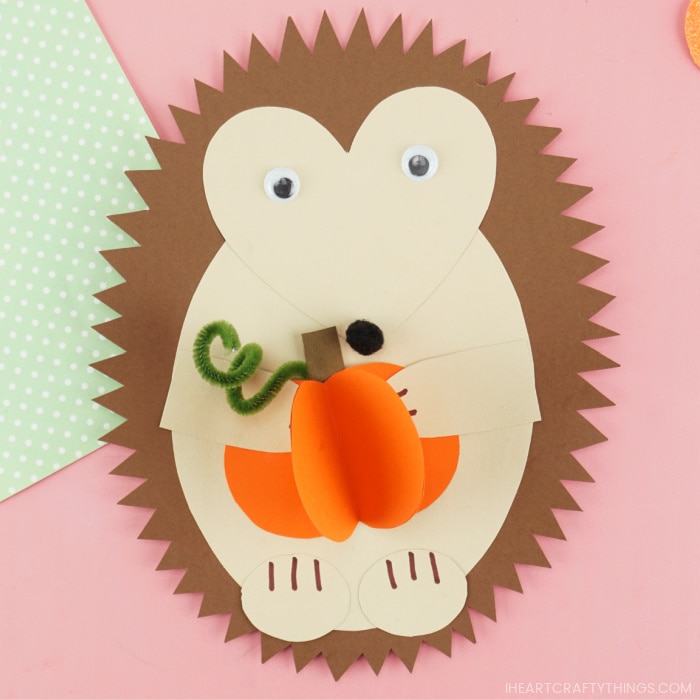 Paper hedgehog craft holding a 3D paper pumpkin laying flat on a pink background with the corner of a green polkadot piece of scrapbook paper showing in the top right corner.
