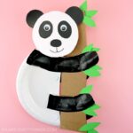 Square image of paper plate panda bear craft holding onto cardboard tree trunk with leaves.