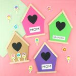 Four Mother's Day birdhouse cards in pink, green, purple and tan laying flat on a pink background with flower stickers scattered around