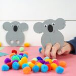 square image of someone playing with a koala finger puppet with scattered pom poms on pink table and white background