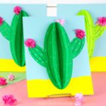 three paper cactus crafts propped up on pink table with white shiplap background