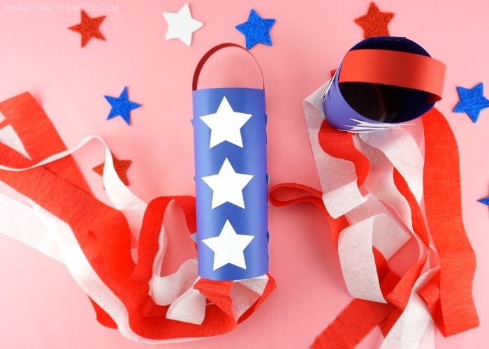 One patriotic windsock laying flat and one standing up on a pink background with glitter star stickers scattered around.