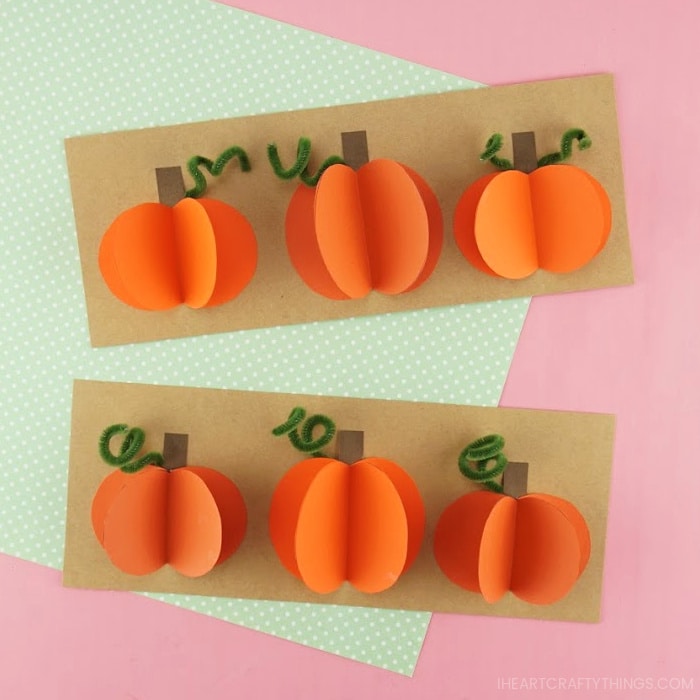 Two 3D paper pumpkin crafts laying one on top of the other on a pink background with a piece of green polkadot scrapbook paper laying behind it.