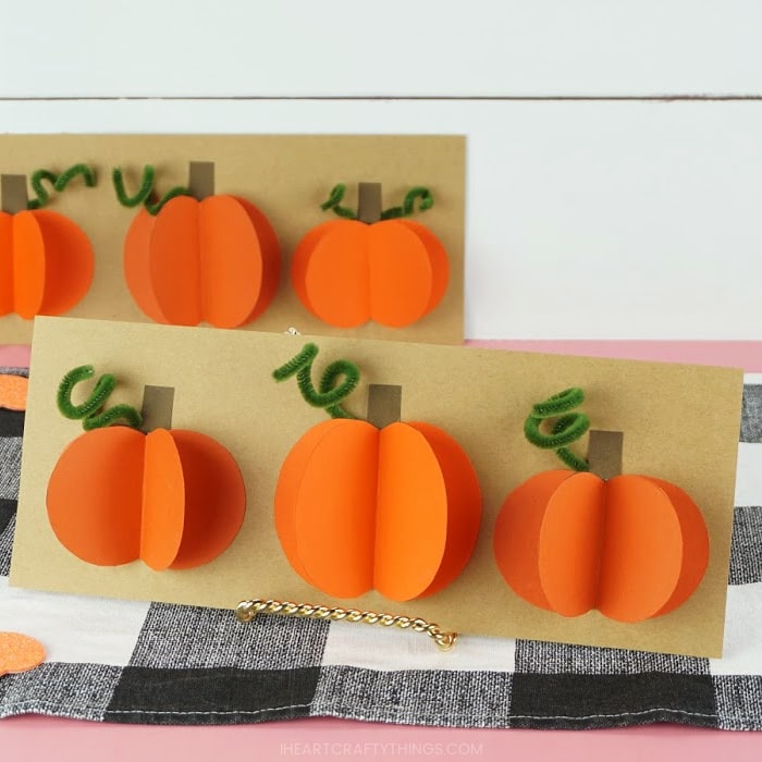 Square image of one paper pumpkin craft up close and in focus propped up and laying on a buffalo plaid placemat, and one in the background out of focus.