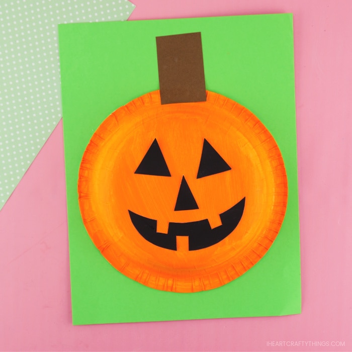 Paper bowl pumpkin craft glued onto green cardstock paper, laying on pink background with a sheet of green polkadot scrapbook paper in the corner.
