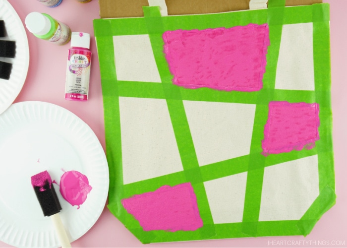 Close up image of canvas bag with three of the taped off shapes painted with pink acrylic paint. Paper plate with pink paint and spongebrush laying in the bottom left corner.