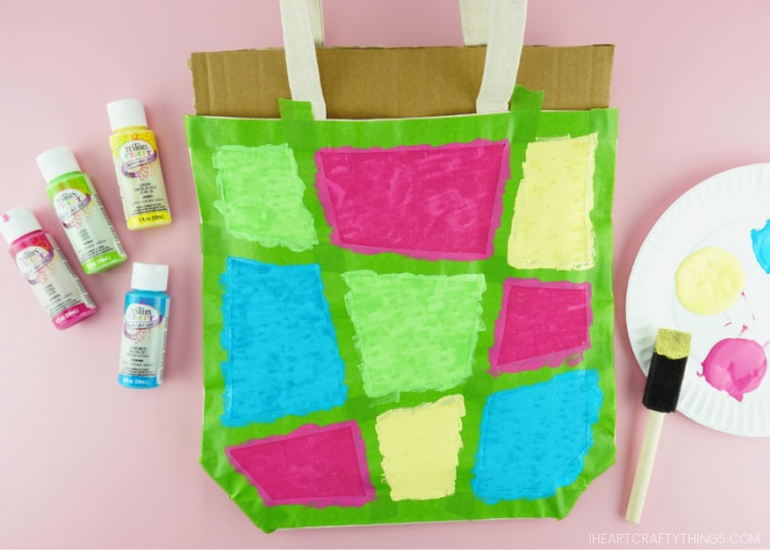 How the canvas bag looks after painted with pink, blue, green and yellow paint waiting to dry. Testors paint laying on left of bag and paper plate paint palette and sponge brush laying on right of painted bag.