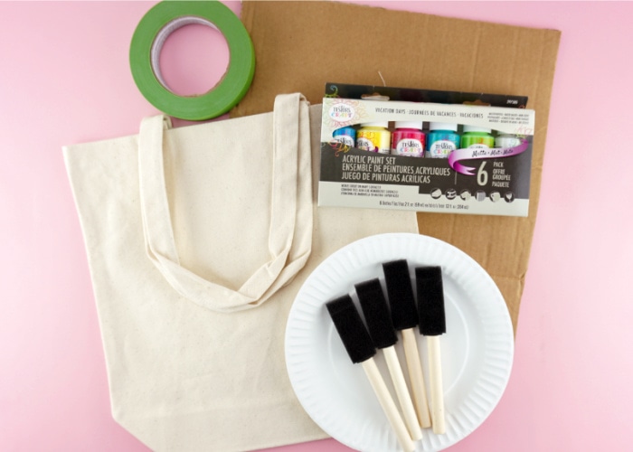 Materials needed to make the painted canvas bag laid out: canvas bag, cardboard, acrylic paint, painters tape, paper plates and sponge paintbrushes.