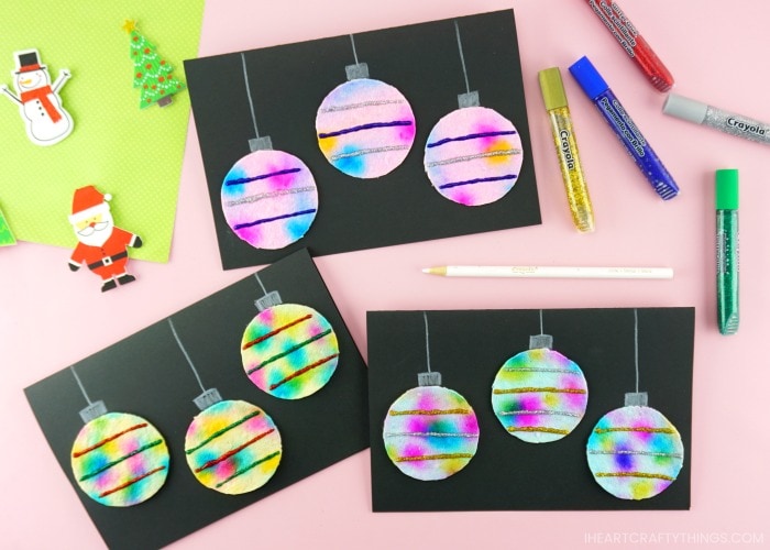 Horizontal image displaying three folded hanging ornaments Christmas cards on a pink table with white colored pencil and glitter glue scattered around the cards.