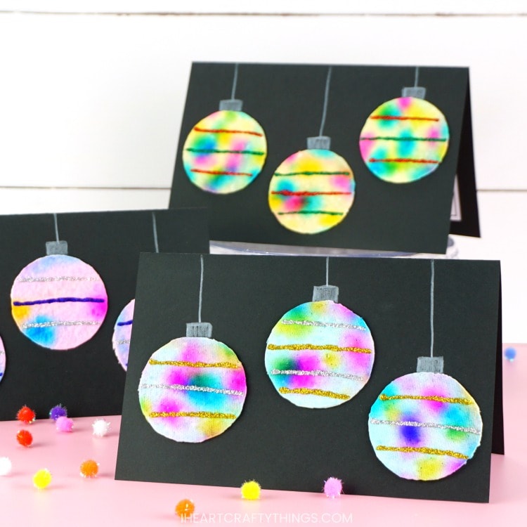 Three DIY hanging ornaments Christmas cards on display on a pink table with a white shiplap background. The card in front is in focus and the ones in back are out of focus.