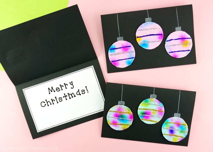 Two hanging ornaments Christmas cards laying flat on a pink background and one laying open with the message "Merry Christmas" on the inside.