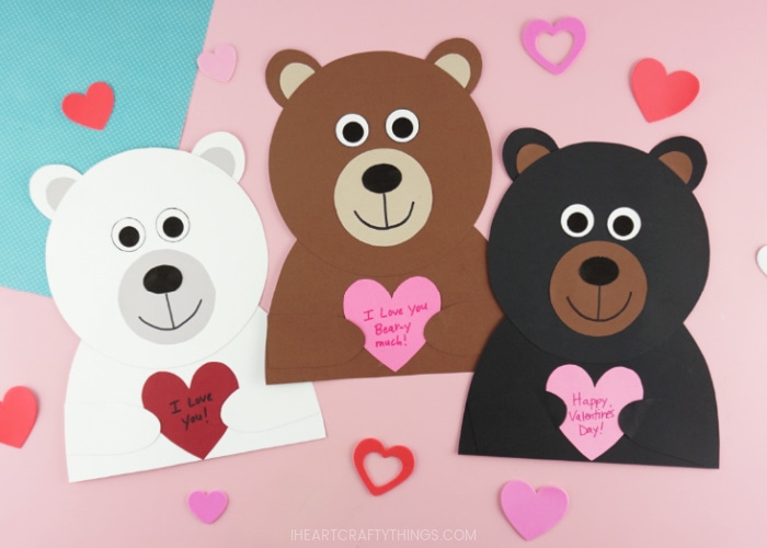 Horizontal image showing a polar bear, brown bear and black bear Valentine craft all laying next to each other on a pink background with heart stickers scattered around them.